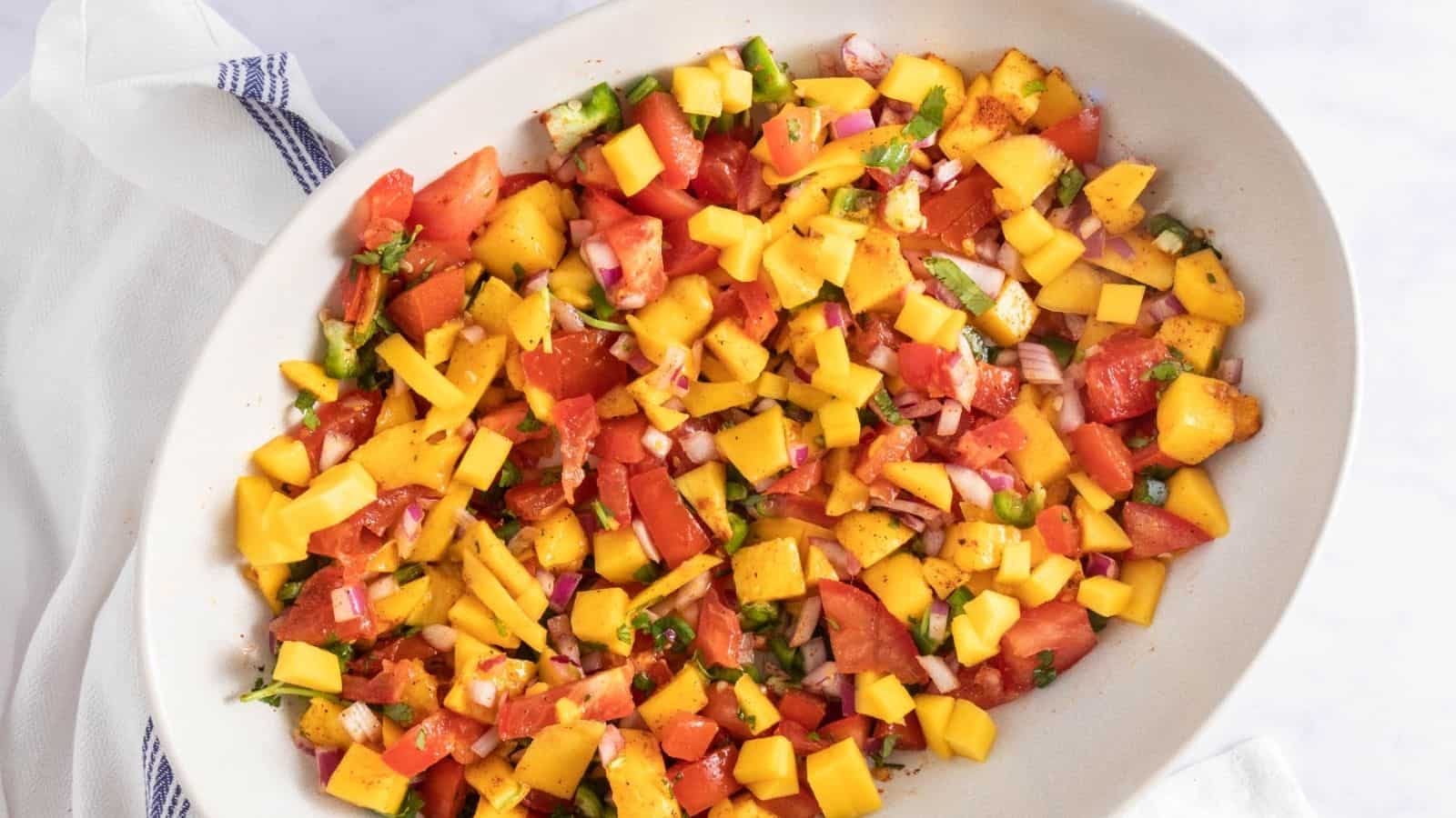A white oval dish filled with a colorful mango salsa containing chopped mango, tomatoes, red onion, jalapeรฑo, and herbs.