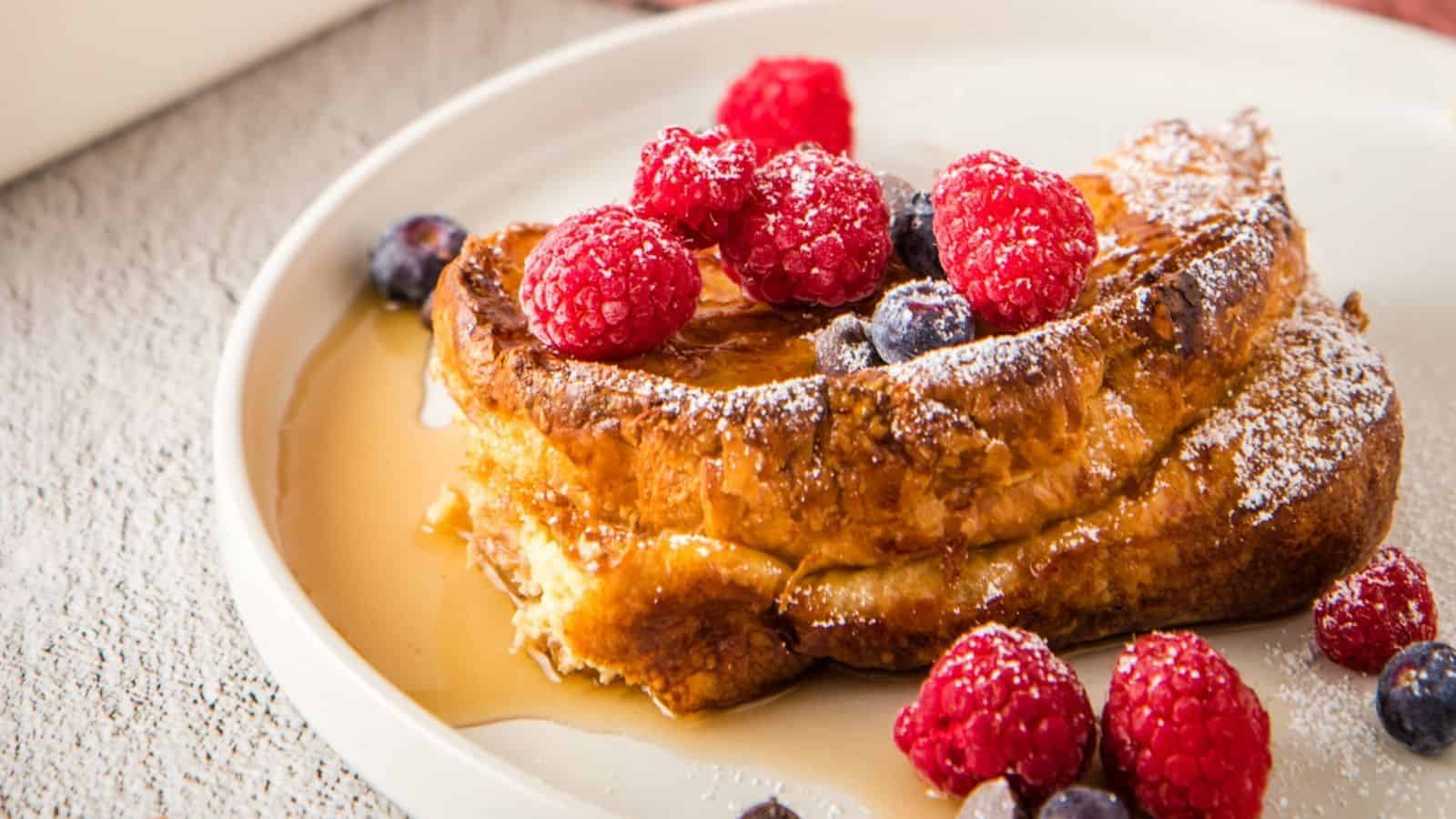 A slice of French toast topped with fresh raspberries, blueberries, powdered sugar, and syrup on a white plate.
