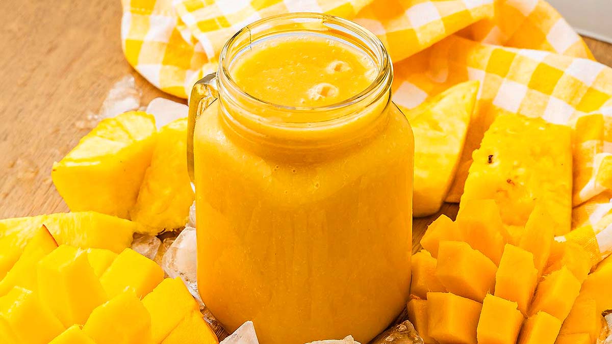 A glass jar filled with yellow smoothie is surrounded by cut pieces of mango and pineapple on a wooden surface with a yellow checkered cloth.