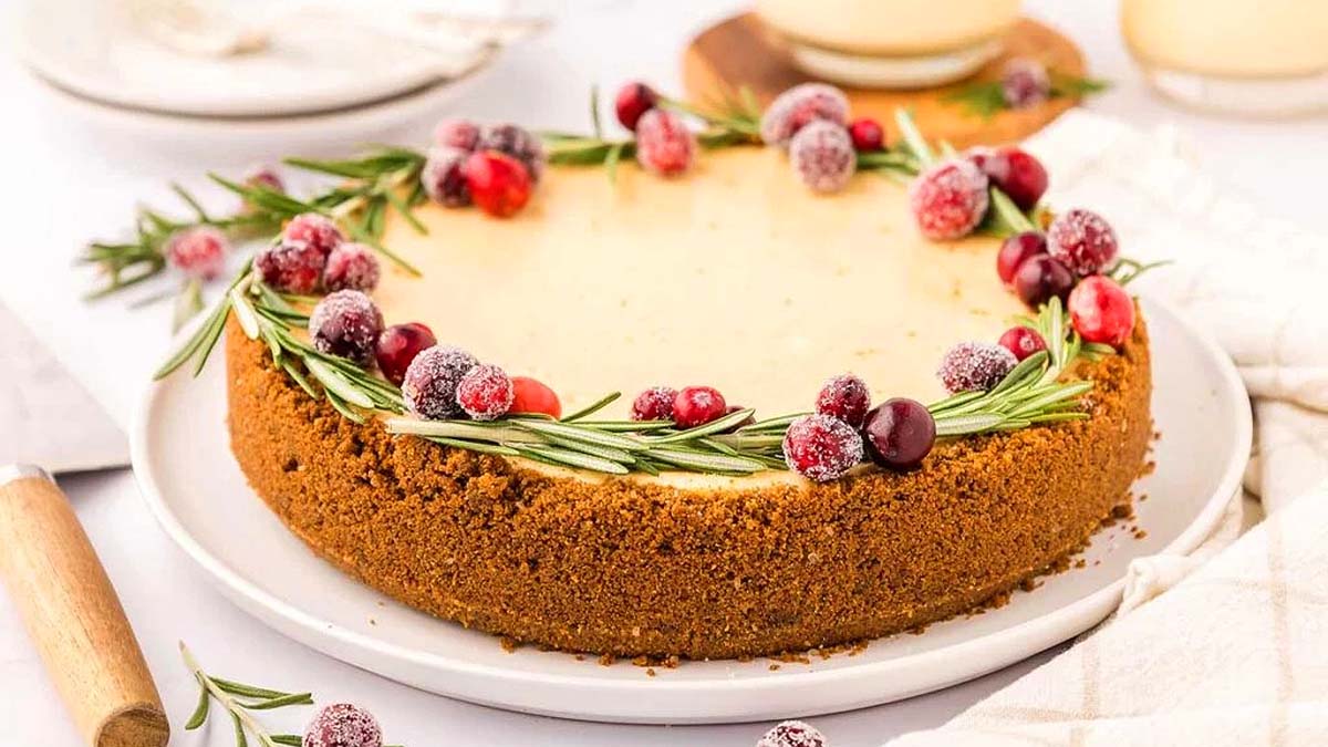 Cheesecake with a graham cracker crust, garnished with sugared cranberries and sprigs of rosemary, served on a white plate.