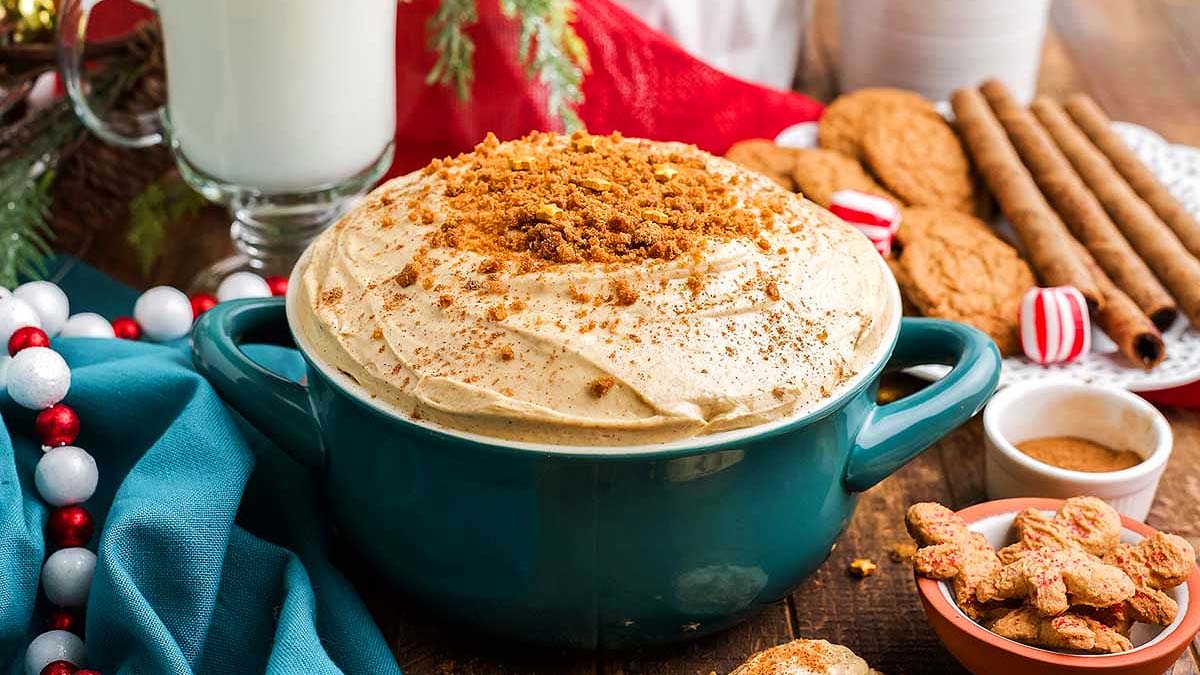 A blue dish filled with a creamy dessert topped with crumbled cookies, surrounded by milk, cookies, spices, and festive decorations on a wooden table.