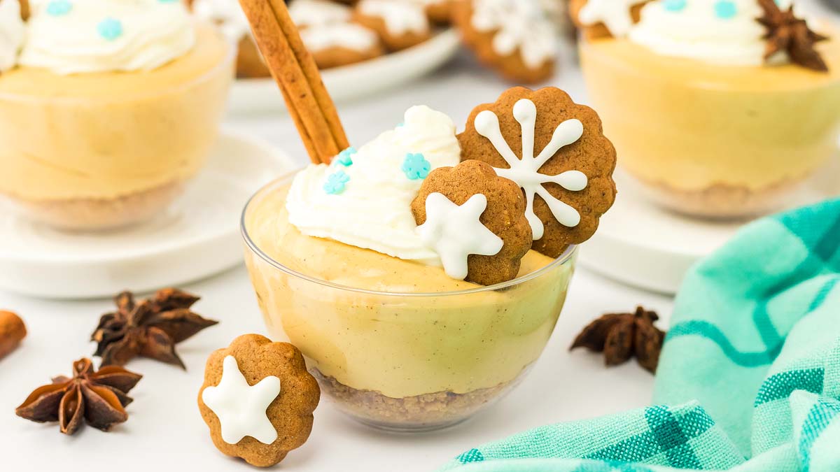 Glass bowl filled with creamy dessert, topped with whipped cream, two gingerbread cookies with white icing, blue sprinkles, and a cinnamon stick; star anise is nearby.