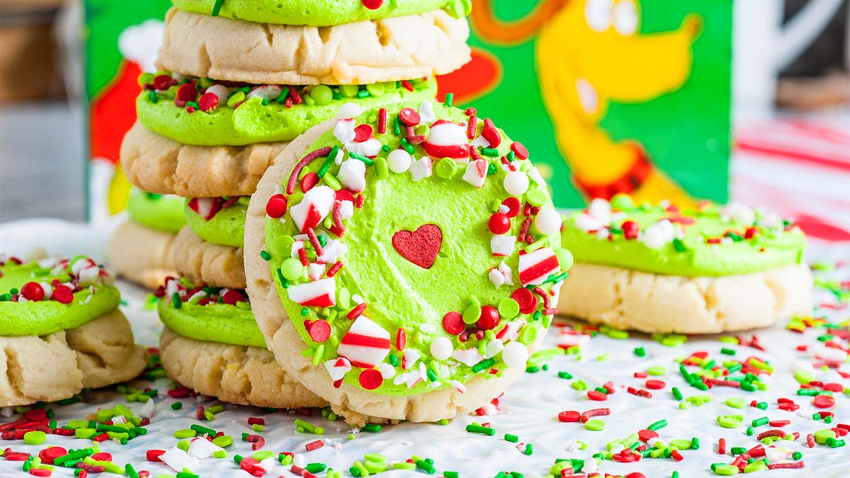 A stack of thick sugar cookies topped with green frosting and colorful holiday sprinkles, with a small red heart in the center, on a festive surface.