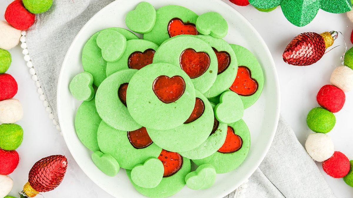 A white plate holds green cookies with heart-shaped red centers, surrounded by festive red, green, and white decorations.