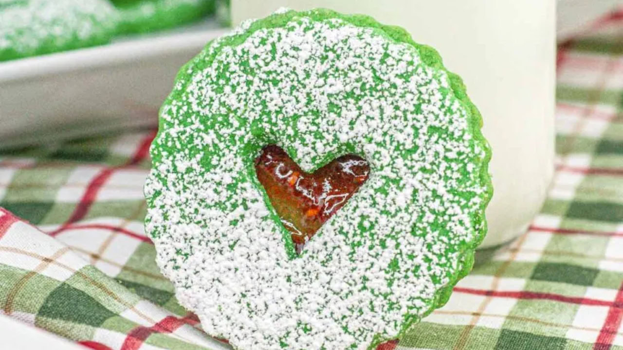 A green round cookie dusted with powdered sugar, featuring a heart-shaped cutout revealing red jam, is displayed upright on a plaid cloth.