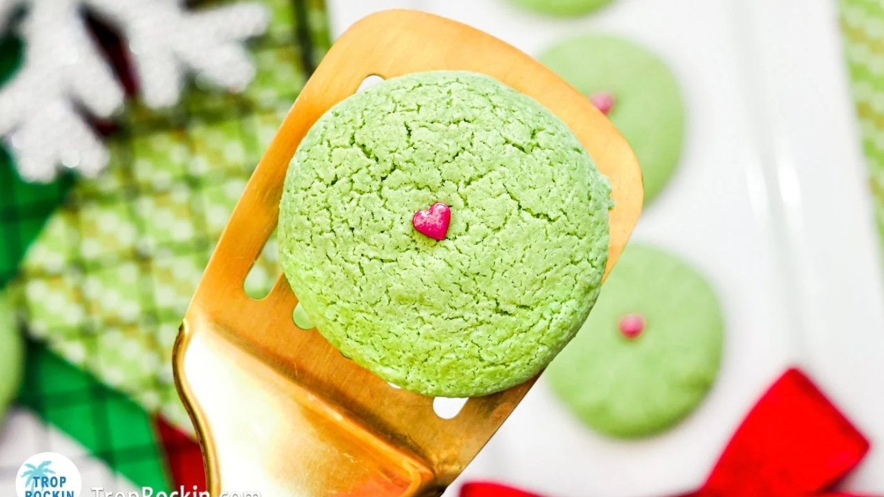 A green cookie with a small pink heart decoration is held on a gold spatula, with more green cookies and holiday decorations in the background.