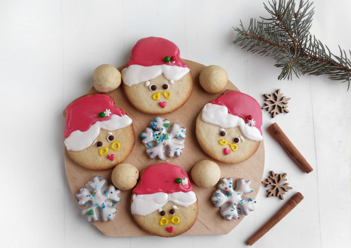 A round wooden board with decorated Christmas cookies shaped like Santa faces and snowflakes, surrounded by cinnamon sticks, star anise, and a pine branch.