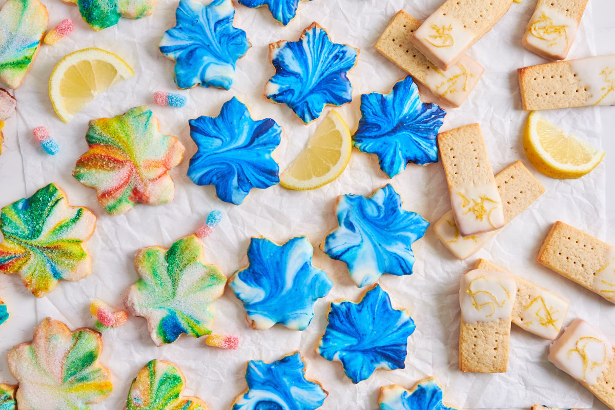 Assorted decorated cookies, including blue leaf-shaped cookies, rainbow sugar cookies, and iced shortbread, arranged on parchment paper with lemon slices.
