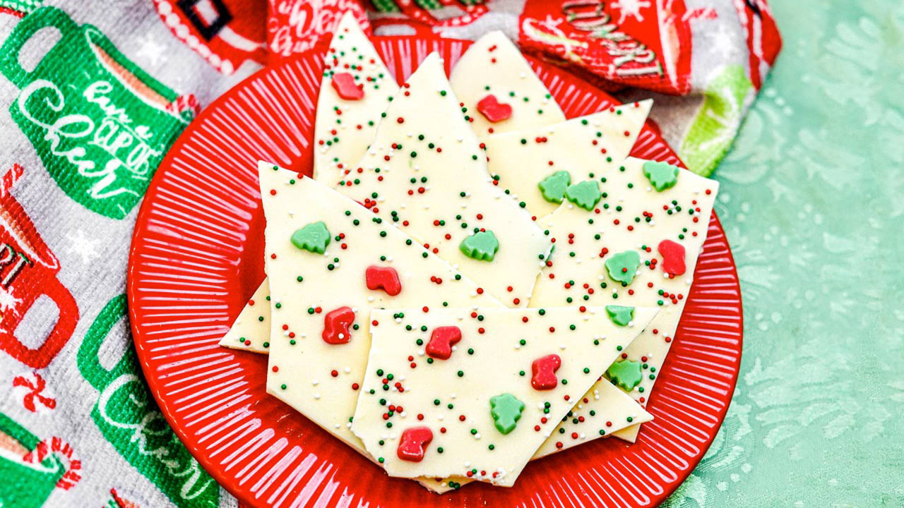 A red plate holds pieces of white chocolate bark topped with red and green sprinkles and Christmas tree-shaped candies, set on a green surface with holiday-themed fabric nearby.