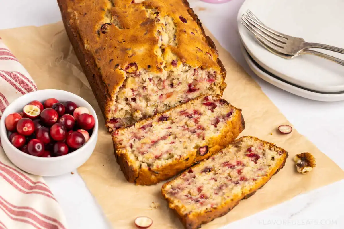 A loaf of cranberry bread with two slices cut, placed on parchment paper next to a bowl of fresh cranberries and a stack of plates with a fork.