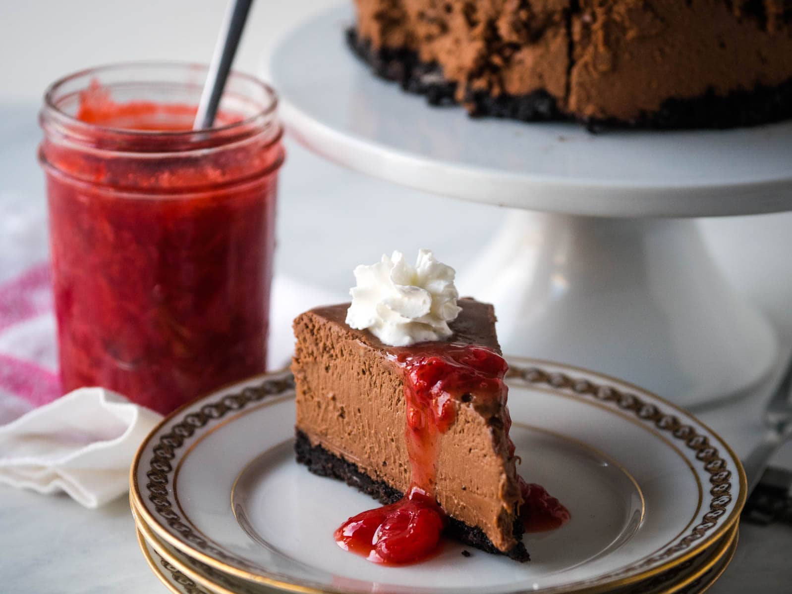 A slice of chocolate cheesecake with whipped cream and strawberry sauce on a plate, next to a jar of strawberry sauce and a cake stand holding the rest of the cheesecake.