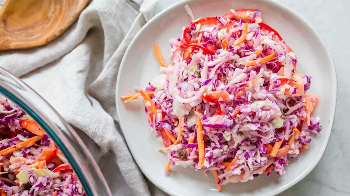A plate of coleslaw with shredded red and green cabbage, carrots, and red bell peppers on a white surface.