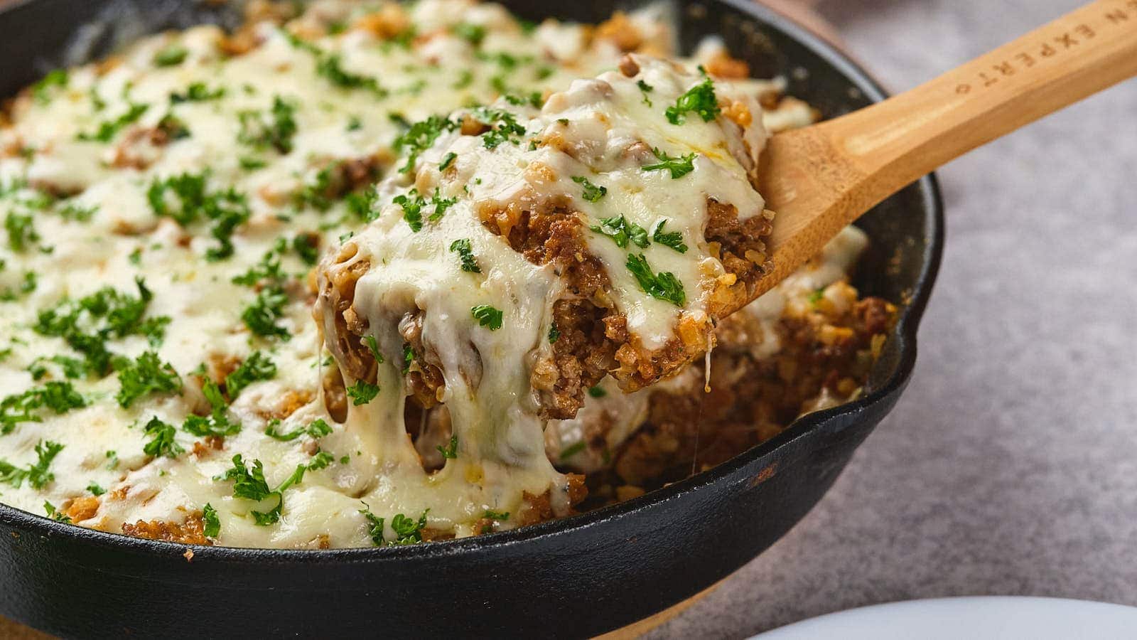 A wooden spoon lifts a serving of cheesy casserole with ground meat and herbs from a black cast iron skillet.