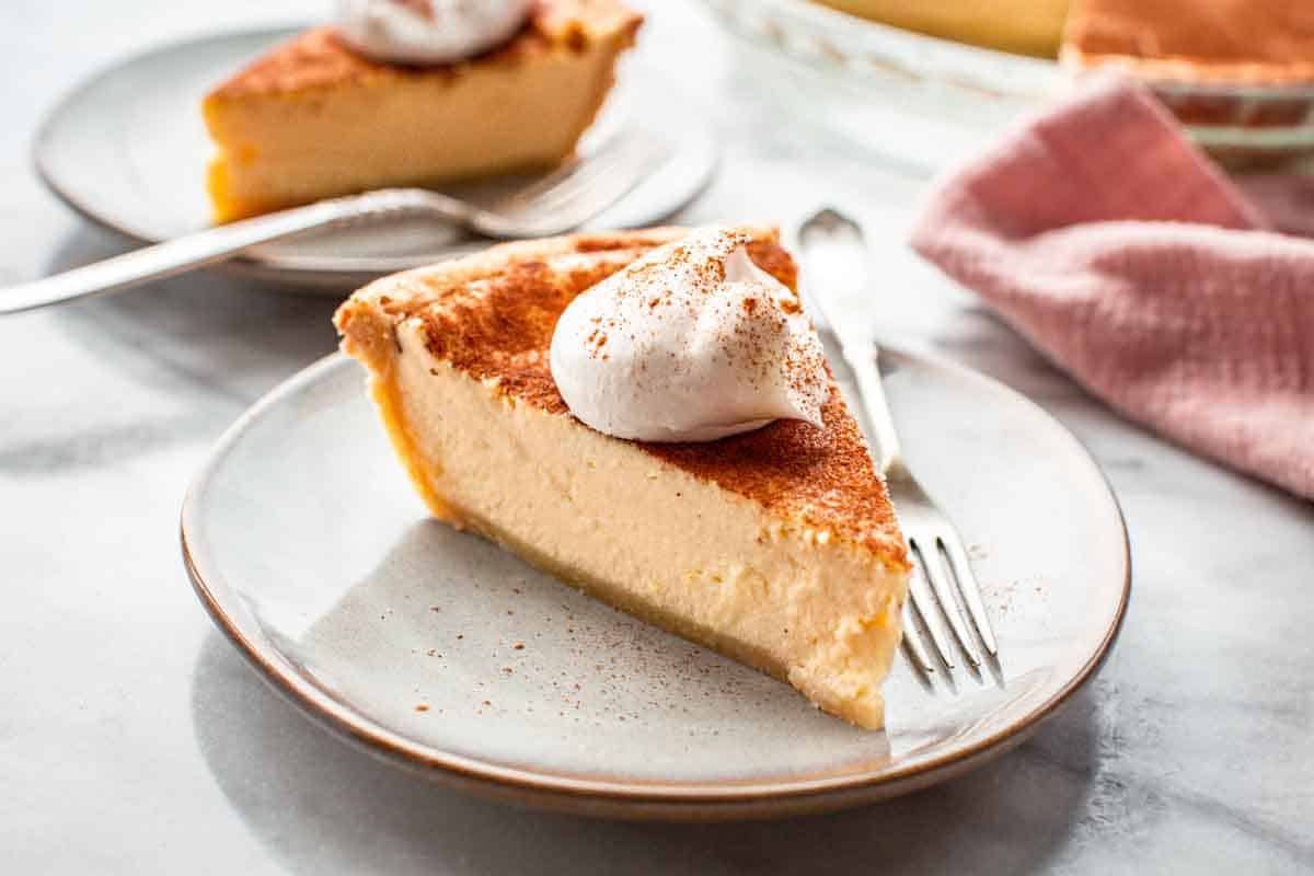 A slice of custard pie topped with whipped cream and a sprinkle of cinnamon on a plate with a fork; another slice and a pink napkin are in the background.