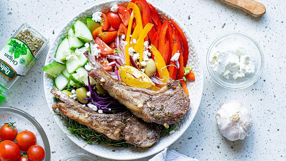 A white bowl with grilled lamb chops, cucumber, cherry tomatoes, red onion, bell pepper, olives, shredded cabbage, and crumbled feta on a light surface.