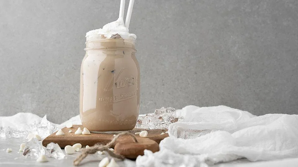 A mason jar filled with a creamy iced beverage topped with whipped cream and two straws, placed on a wooden board with scattered white chocolate chips and ice cubes.
