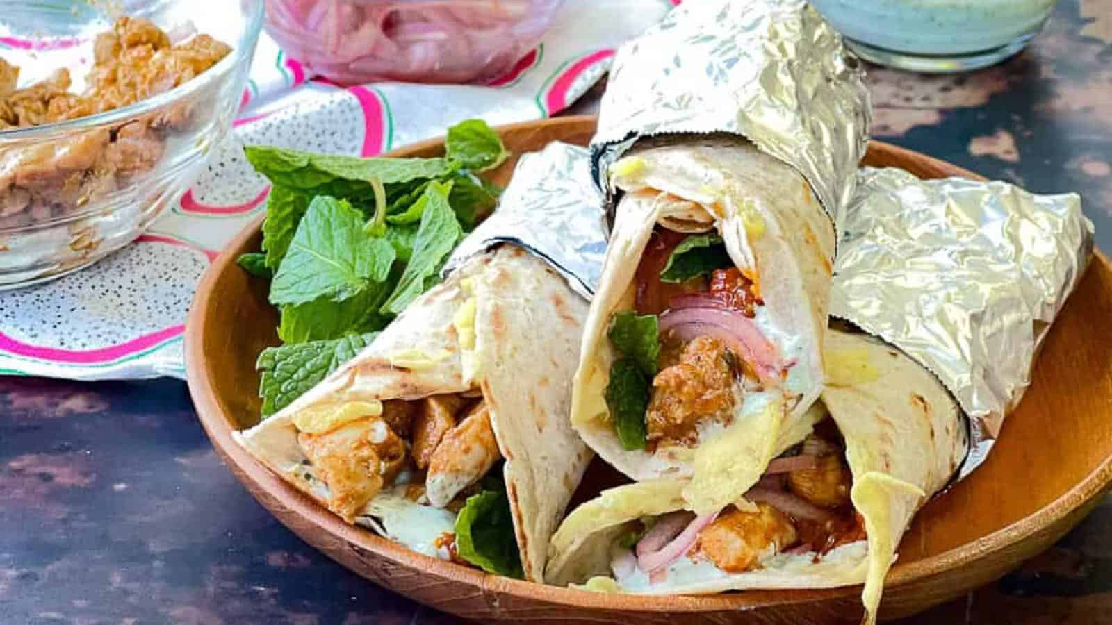 Three chicken wraps filled with greens, onions, and sauce are partially wrapped in foil and placed on a wooden plate.