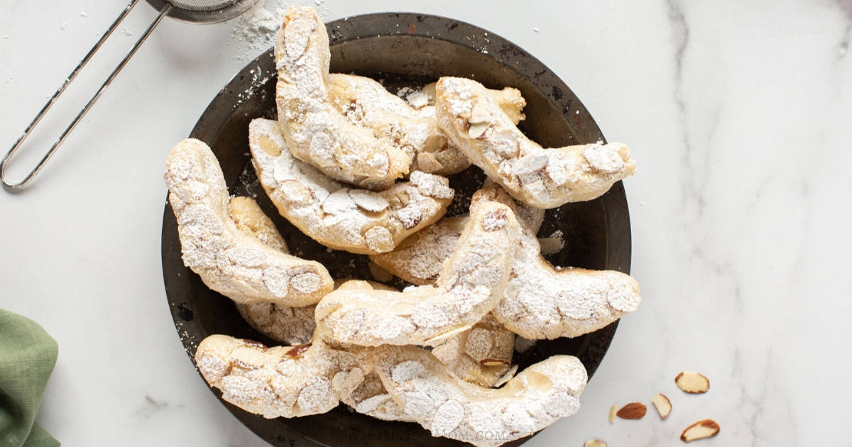 A plate of crescent-shaped almond cookies dusted with powdered sugar, with sliced almonds and a sifter on the side.