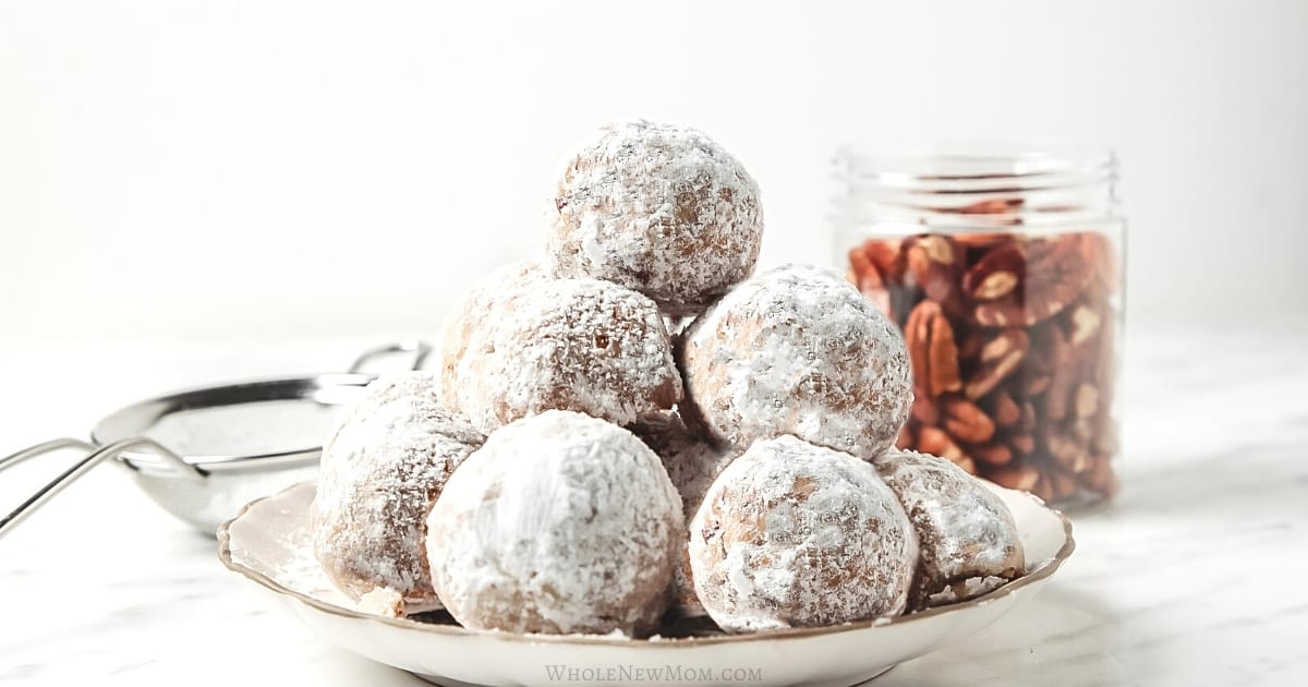 A plate of powdered sugar-coated cookies is stacked in front of a jar filled with pecans on a white surface.