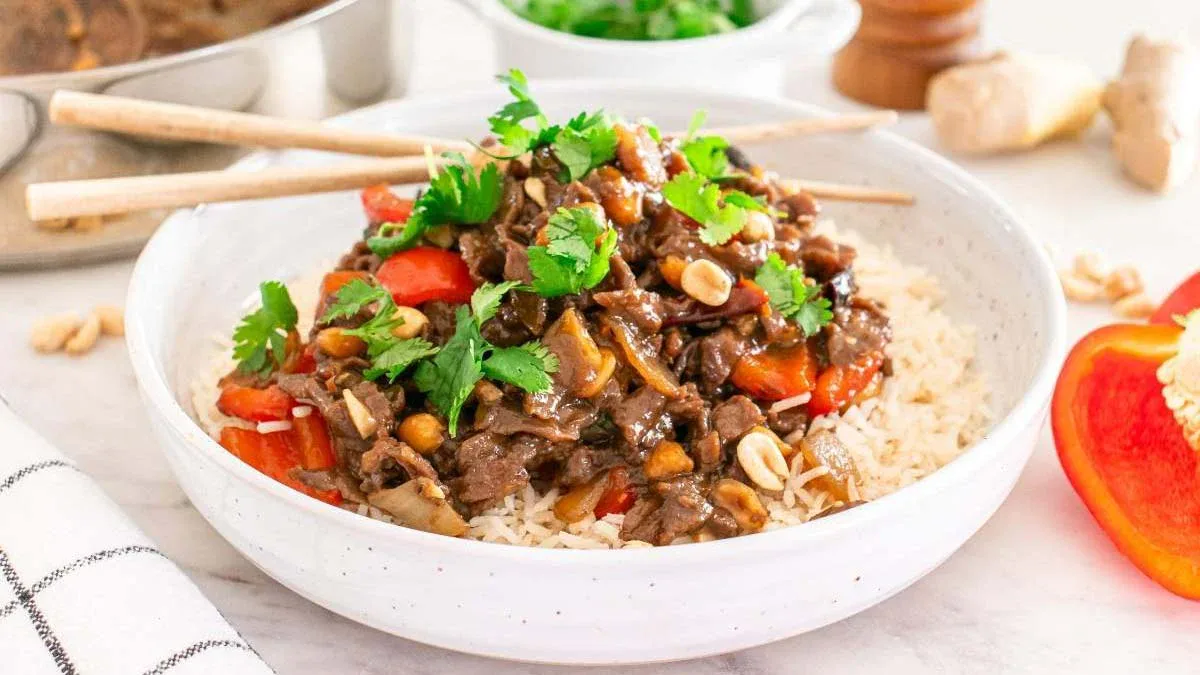 A bowl of white rice topped with stir-fried beef, red bell peppers, peanuts, cilantro, and sauce, with chopsticks resting on the bowl.