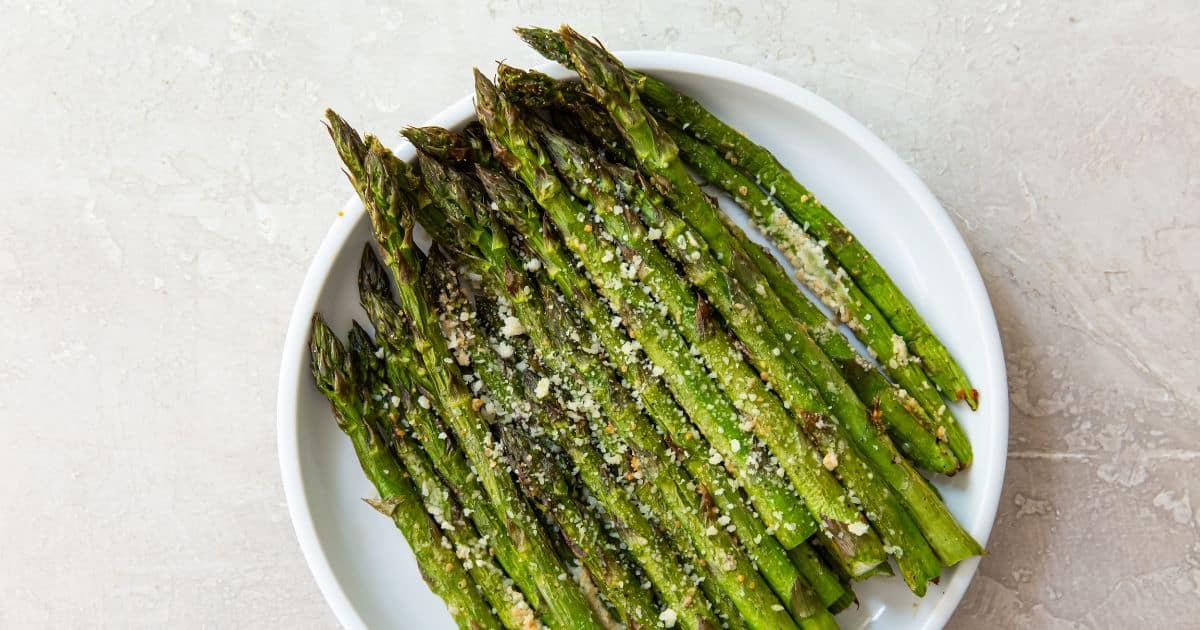 A white plate with roasted asparagus spears sprinkled with grated cheese on a light surface.