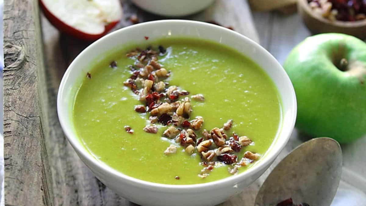 A bowl of green soup topped with chopped nuts and dried fruit, with a green apple and a spoon nearby on a rustic wooden surface.