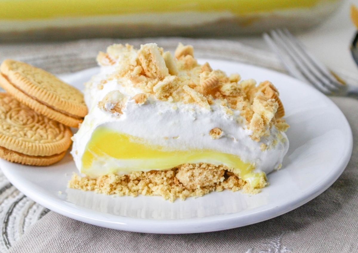 A slice of layered lemon dessert with whipped topping and crumbled cookies on a white plate, served with two whole golden sandwich cookies.