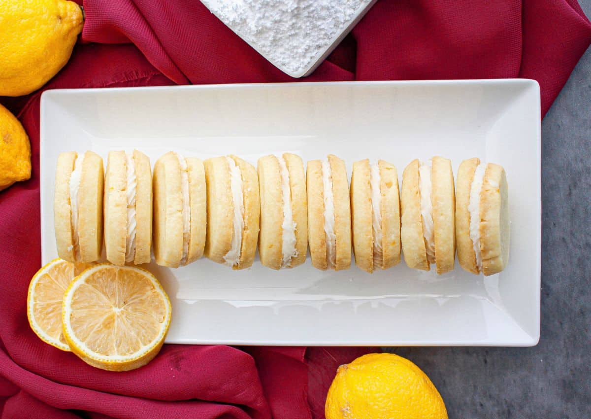 A row of lemon cream sandwich cookies on a rectangular white plate, with lemon slices and whole lemons nearby on a red cloth.