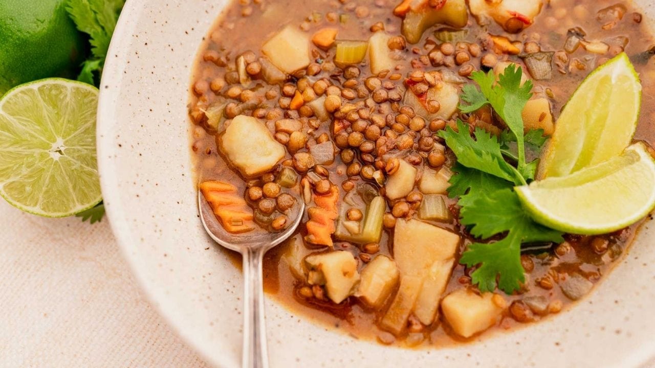 A bowl of lentil soup with diced vegetables, garnished with cilantro and lime wedges, with a spoon resting in the bowl.