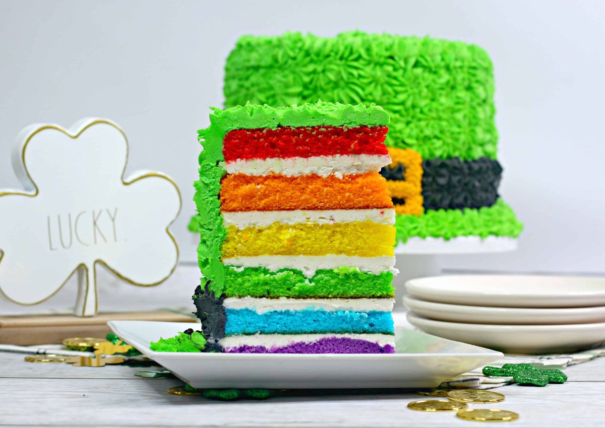 A slice of rainbow layer cake with green icing sits on a plate; the full cake, decorated like a leprechaun hat, is in the background. Gold coins and a shamrock sign are nearby.