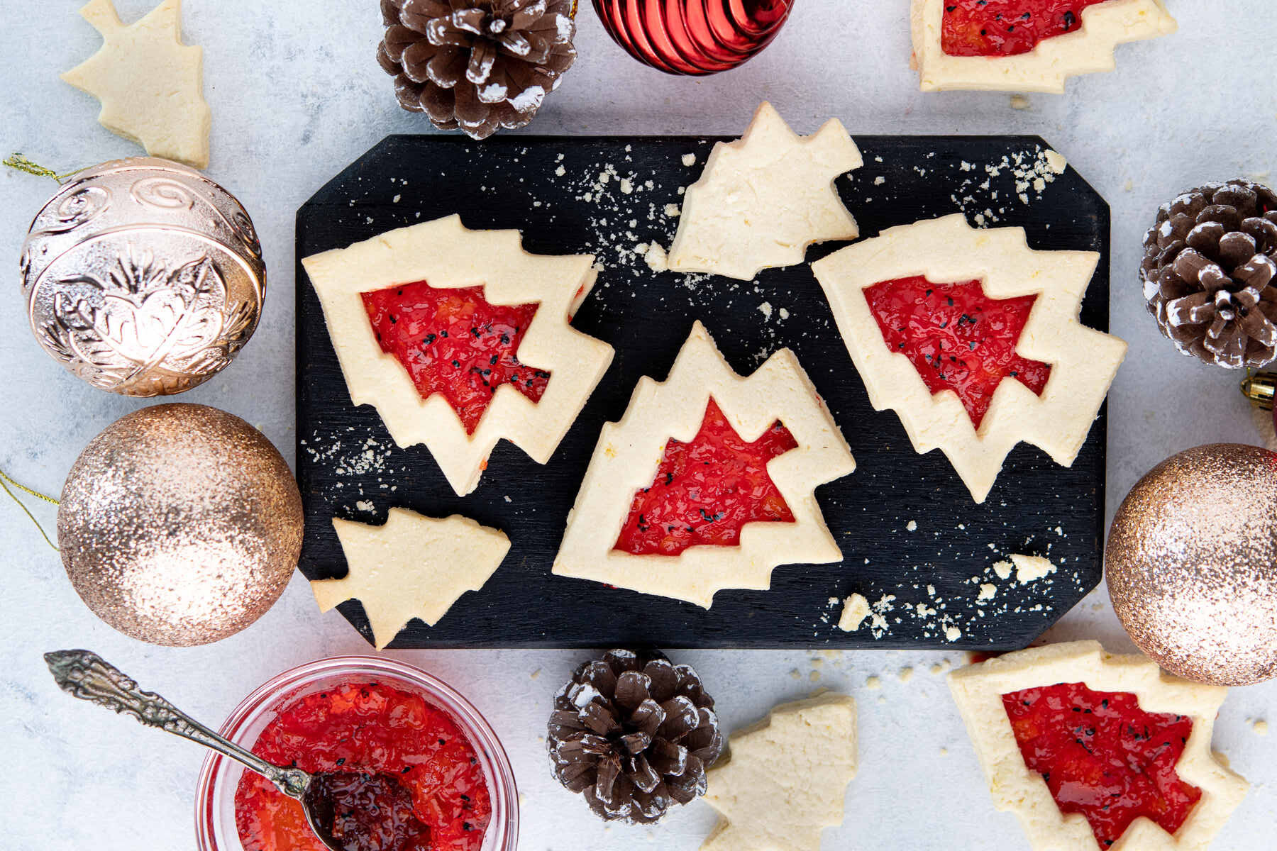 Christmas tree-shaped cookies with red jam centers are arranged on a black slate board, surrounded by pine cones, Christmas ornaments, and a jar of jam with a spoon.