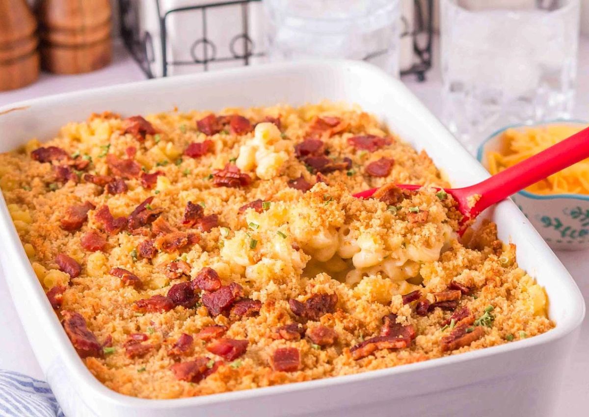 A baking dish filled with macaroni and cheese topped with breadcrumbs and bacon, with a serving spoon scooping some out.