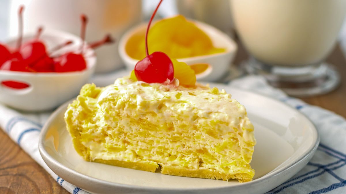 A slice of creamy yellow pie topped with a maraschino cherry and pineapple, served on a white plate with a bowl of cherries and a glass of milk in the background.