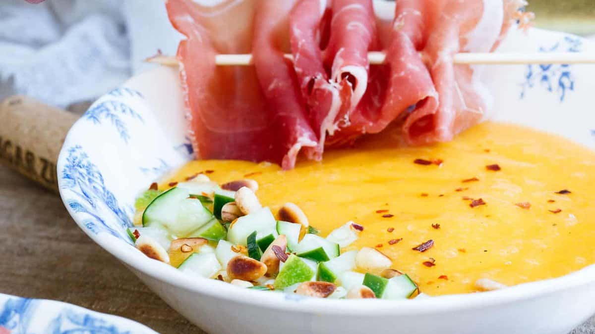 A bowl of yellow soup garnished with diced cucumber, toasted pine nuts, red pepper flakes, and skewered slices of cured meat.