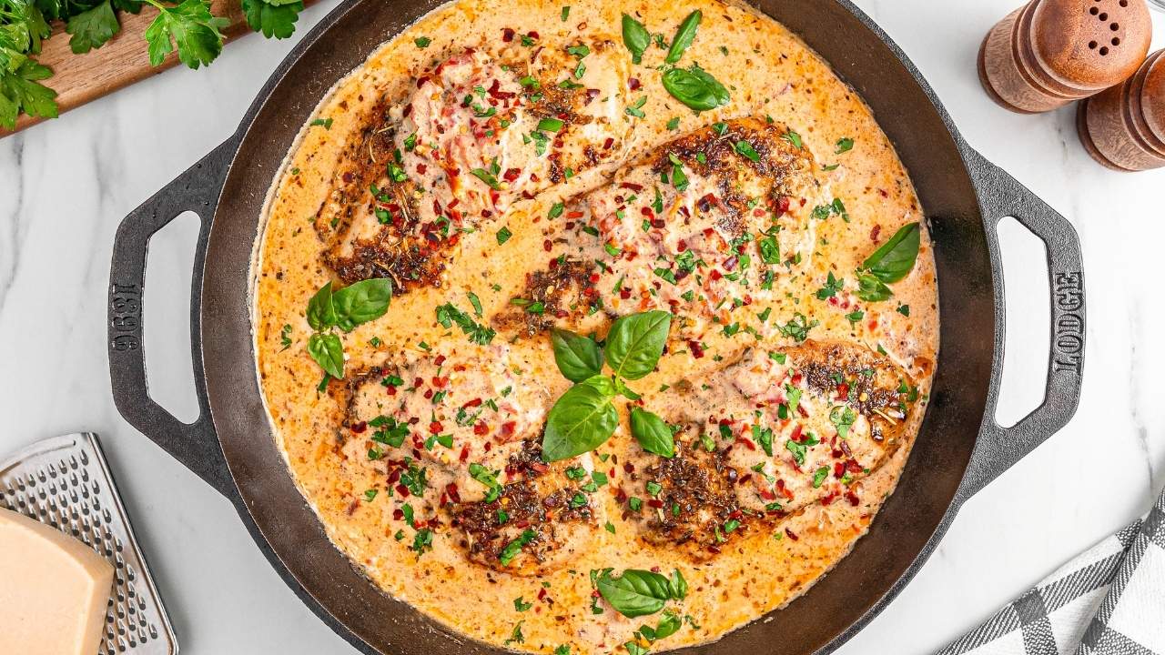 A cast iron skillet filled with creamy sauce and four seasoned, cooked chicken breasts garnished with herbs and red pepper flakes.