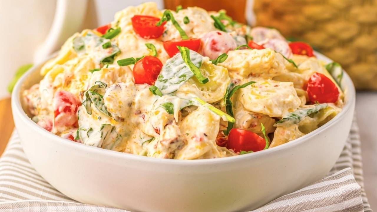 A bowl of creamy potato salad with chopped green onions, cherry tomatoes, and lettuce, served on a striped cloth.