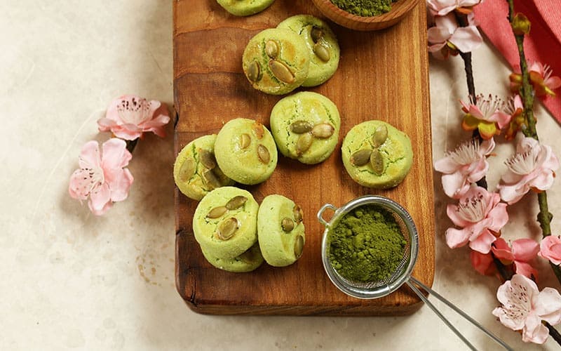 Green cookies topped with pumpkin seeds on a wooden board, with a mesh strainer holding green powder and pink flowers nearby.