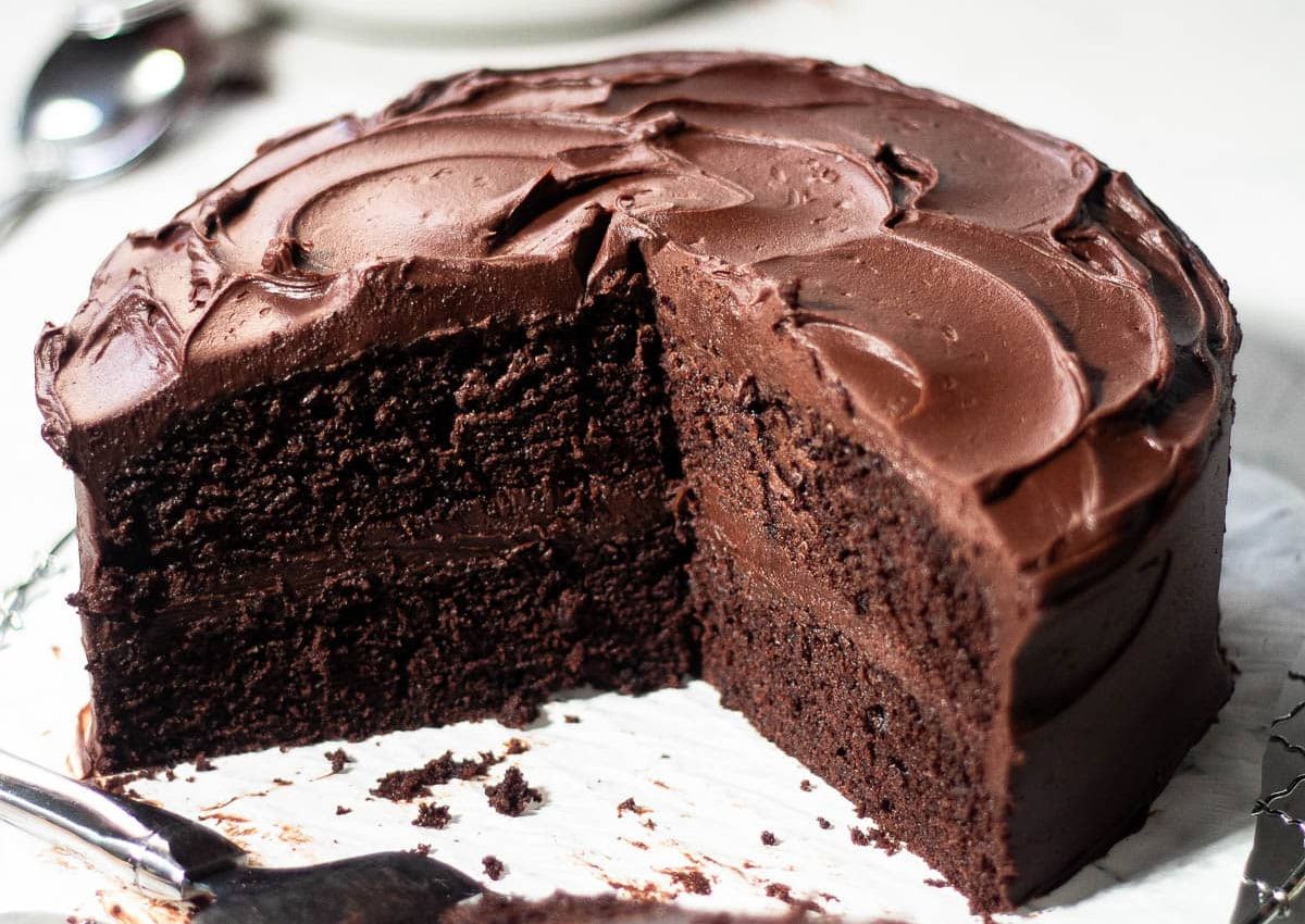 A round, double-layer chocolate cake with chocolate frosting sits on a white surface. A large slice has been cut and is partially removed.