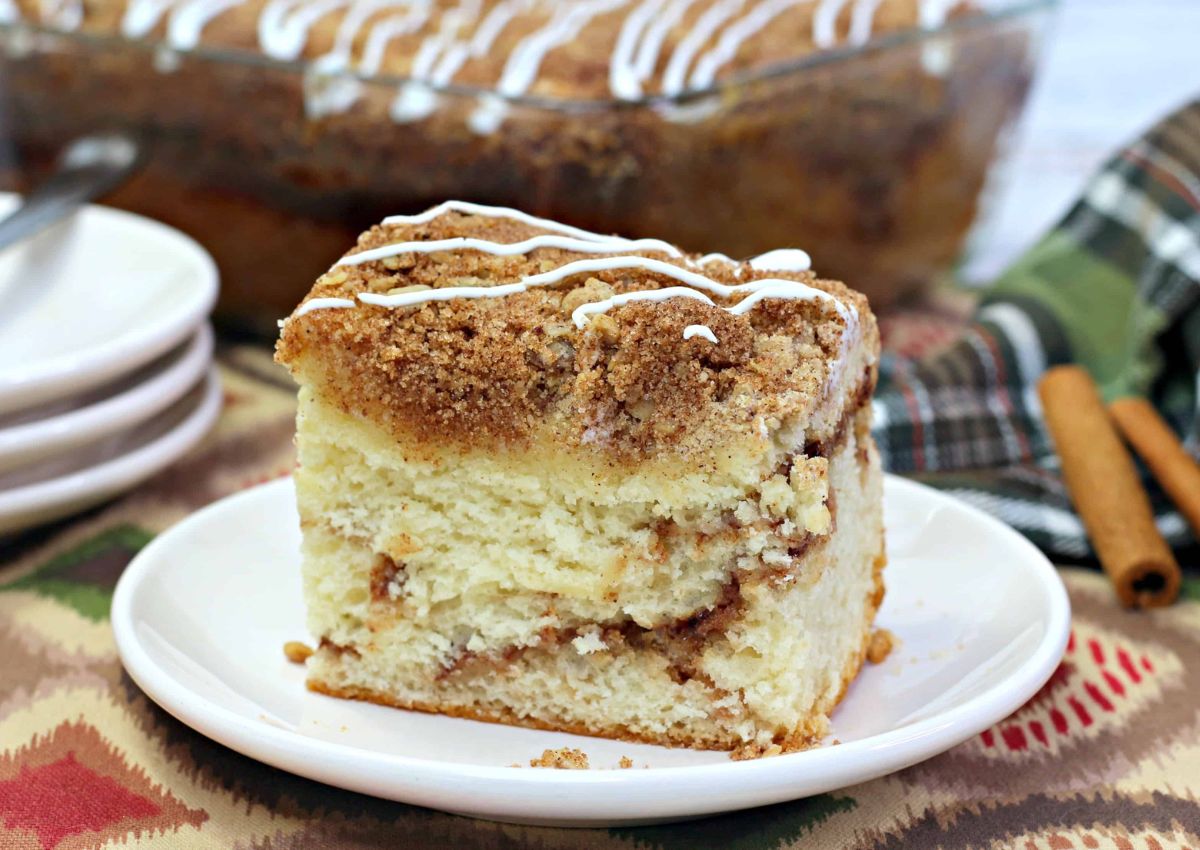 A square slice of coffee cake with cinnamon crumble topping and white icing drizzle sits on a white plate. A baking dish of cake, plates, and cinnamon sticks are in the background.