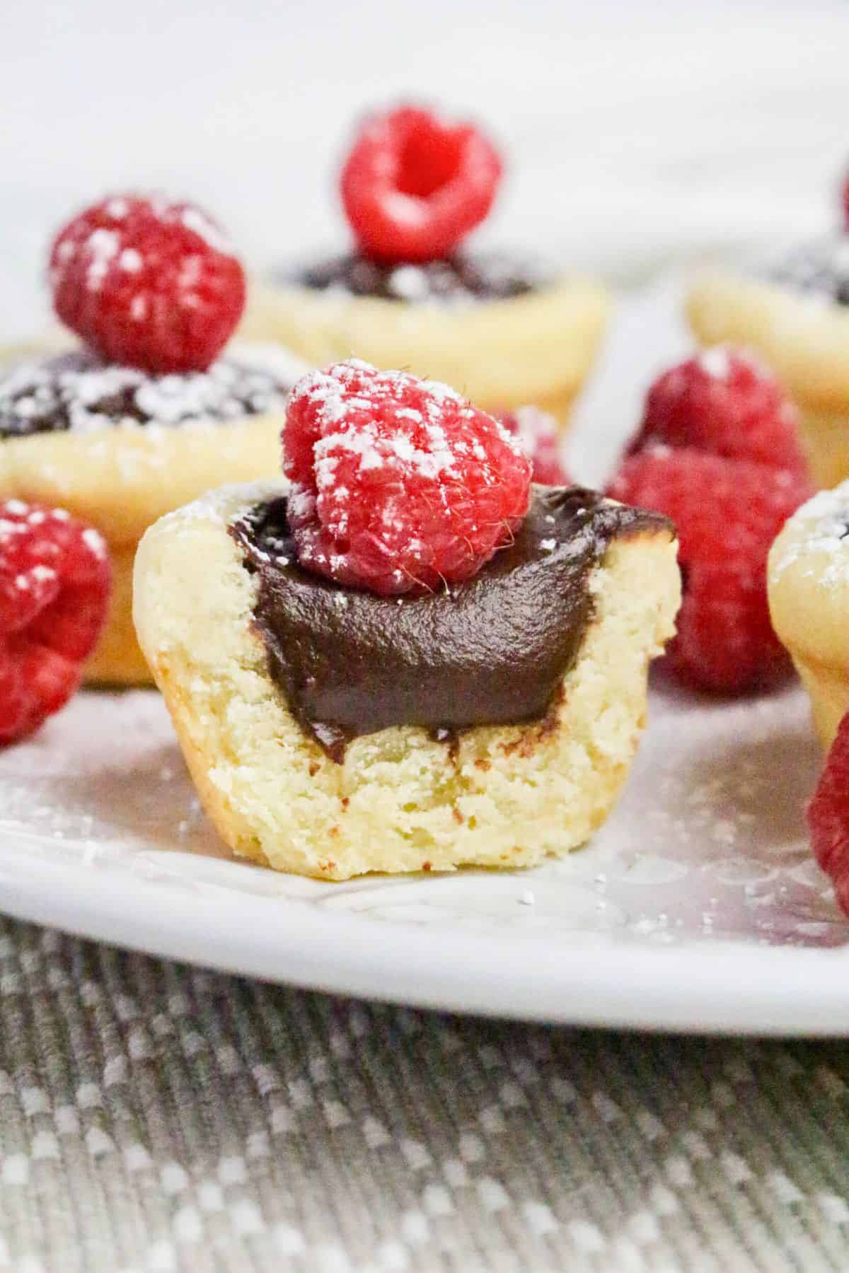 A close-up of a chocolate tart topped with a raspberry and powdered sugar, with a bite taken out, displayed on a white plate with more tarts and raspberries in the background.
