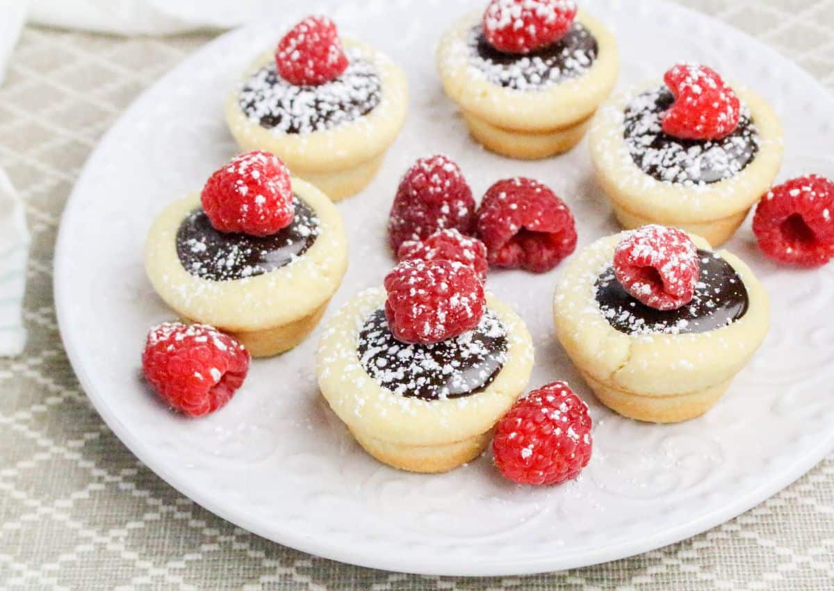 A white plate with six chocolate tartlets topped with raspberries and powdered sugar, surrounded by additional raspberries.