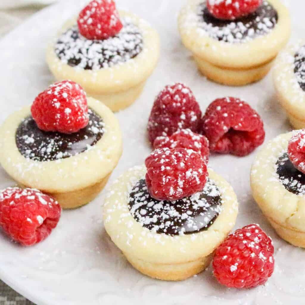 A plate of mini chocolate tarts topped with fresh raspberries and dusted with powdered sugar.
