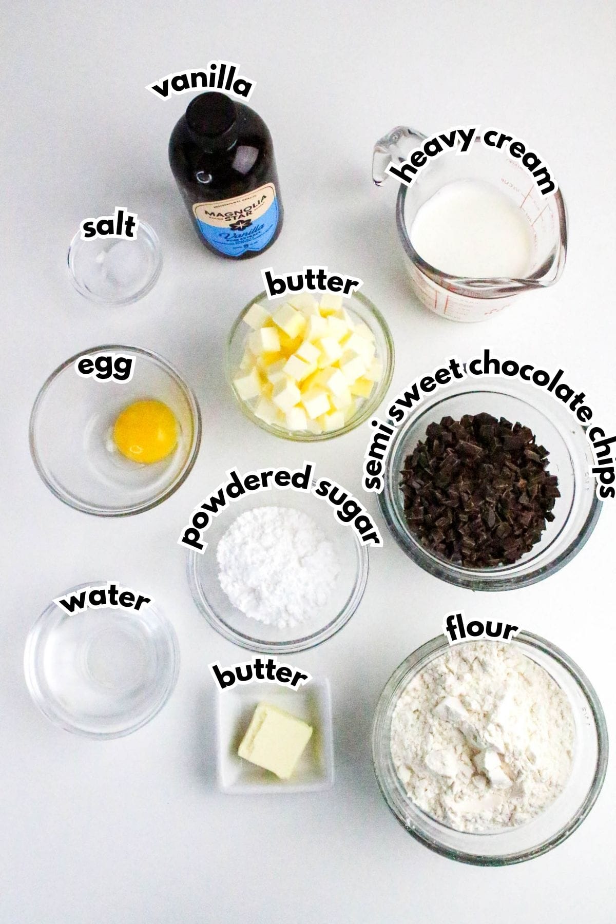 Ingredients for baking are arranged on a white surface, including vanilla, heavy cream, chocolate chips, butter, flour, powdered sugar, salt, egg, and water, each labeled.
