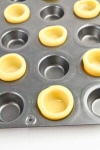 A muffin tin with several cups lined with unbaked dough, forming shallow pastry shells.