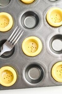 A muffin tin with unbaked tart shells, some pricked with a fork and a metal fork resting inside the tin.