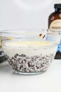 A glass bowl containing chopped chocolate and cream, with a bottle of vanilla extract in the background.