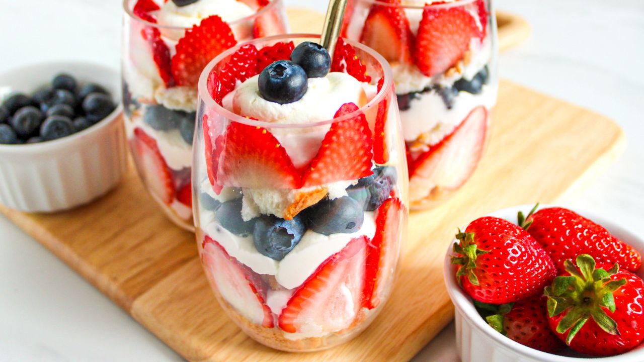 Three glass parfaits layered with sliced strawberries, blueberries, whipped cream, and cake are displayed on a wooden board, with bowls of fresh berries beside them.