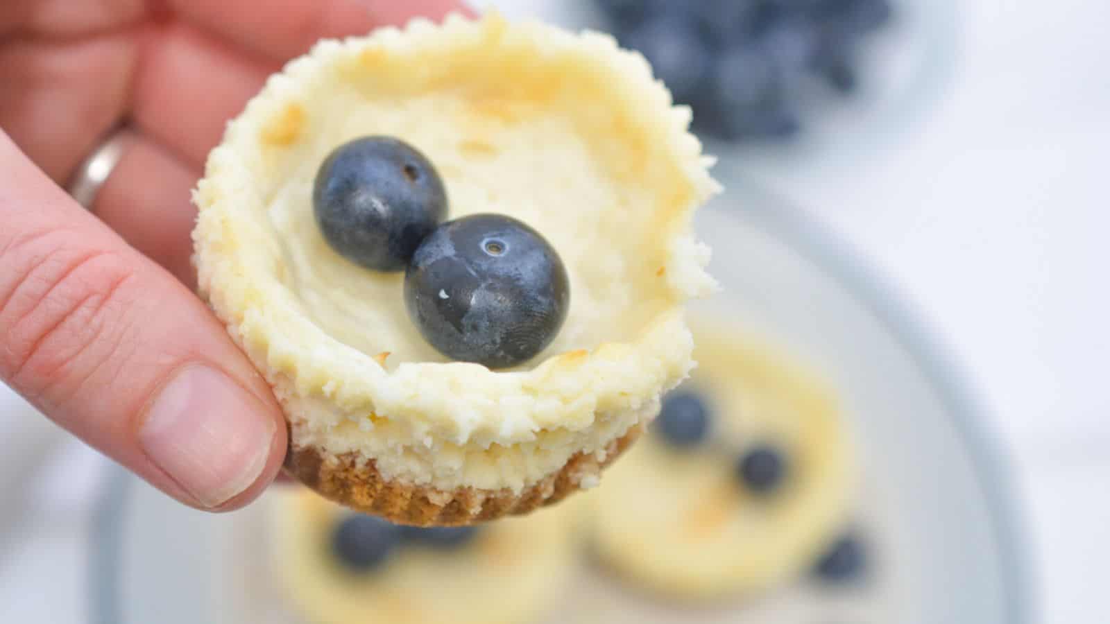 A hand holds a mini cheesecake topped with two blueberries, with more cheesecakes and blueberries on a plate in the background.