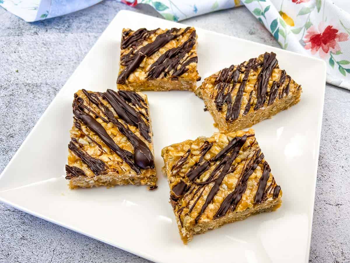Four rectangular oat bars drizzled with chocolate are arranged on a white square plate, with part of a floral napkin visible in the background.