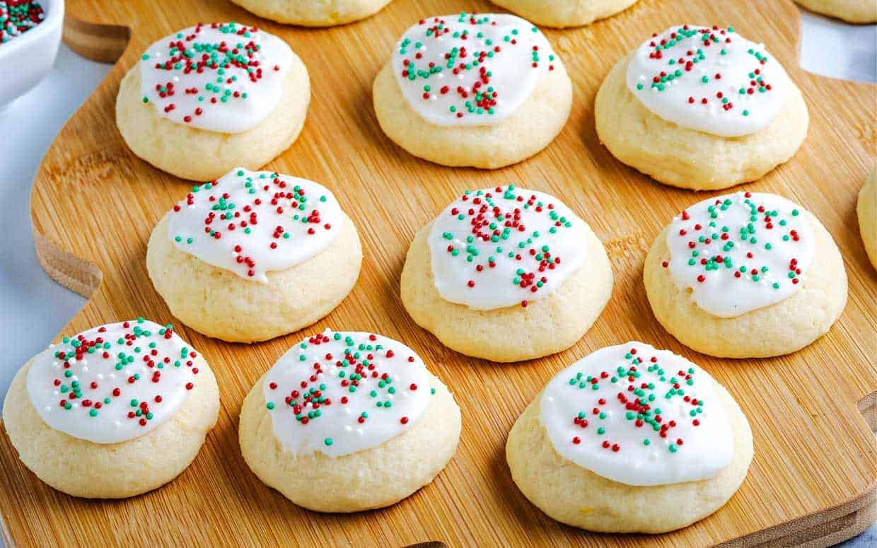 Twelve frosted sugar cookies with red and green sprinkles are arranged on a wooden serving tray.